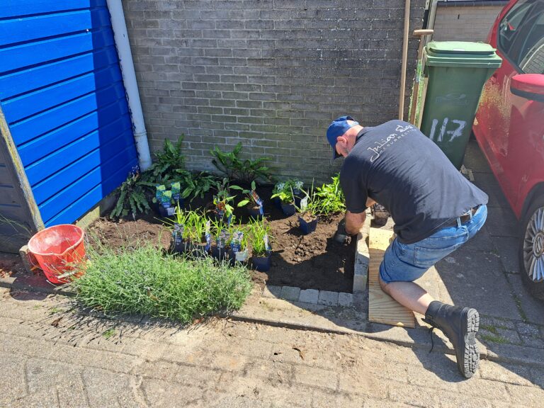 Man plant bloemen in tuin naast blauwe muur en rode auto, met emmer en vuilnisbak op achtergrond.