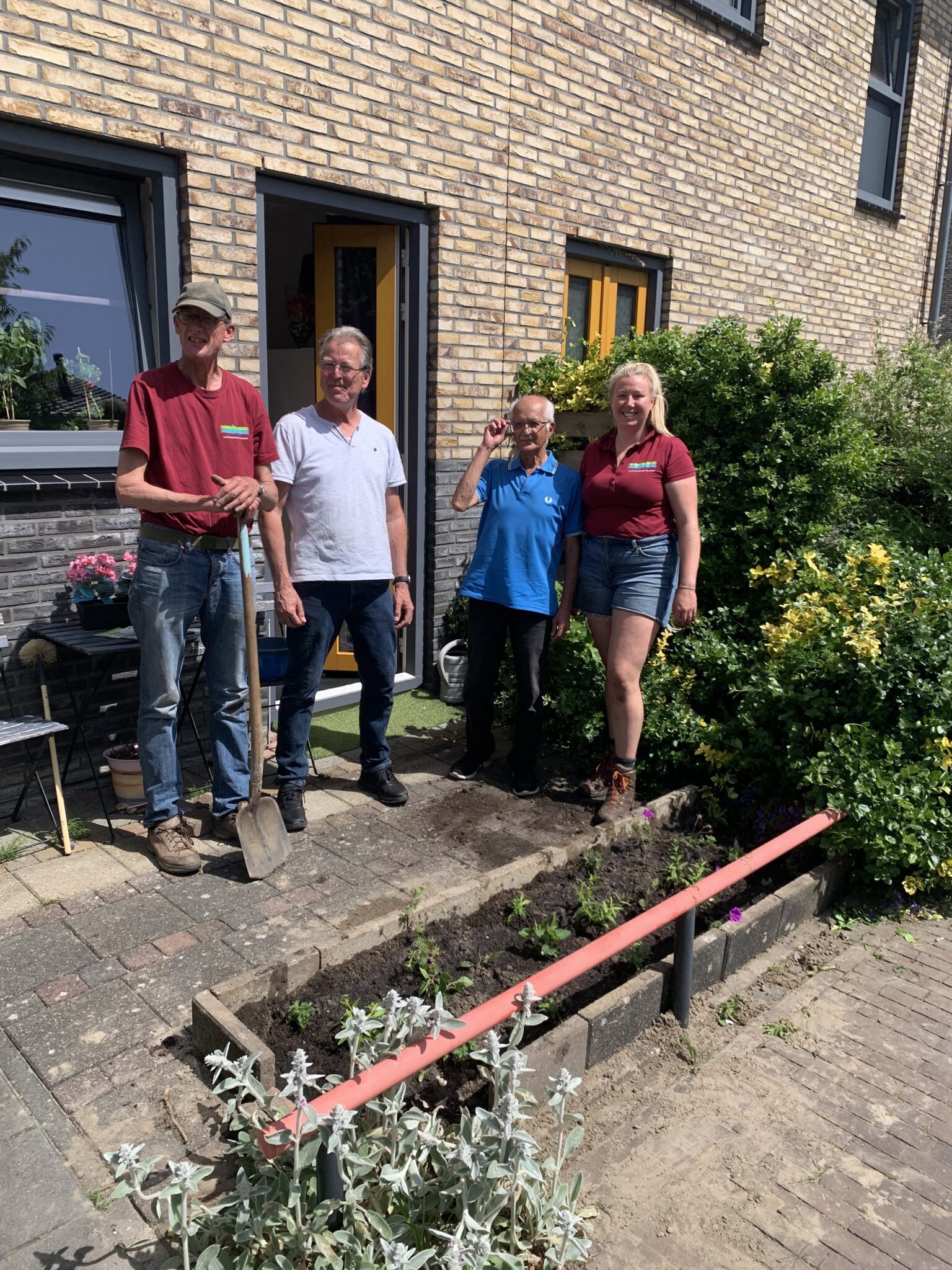 Vier mensen staan voor een huis, naast een tuinperk met jonge planten.
