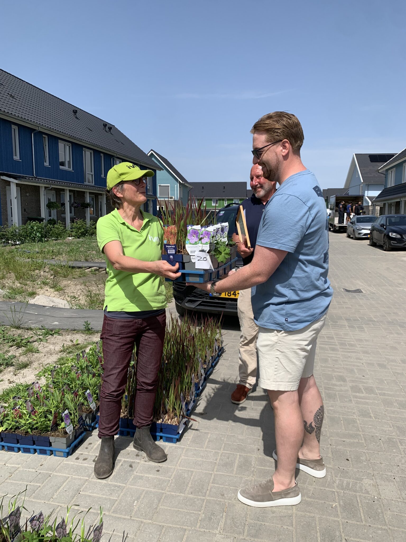 Twee mensen wisselen plantjes uit op een straat met huizen in de achtergrond.