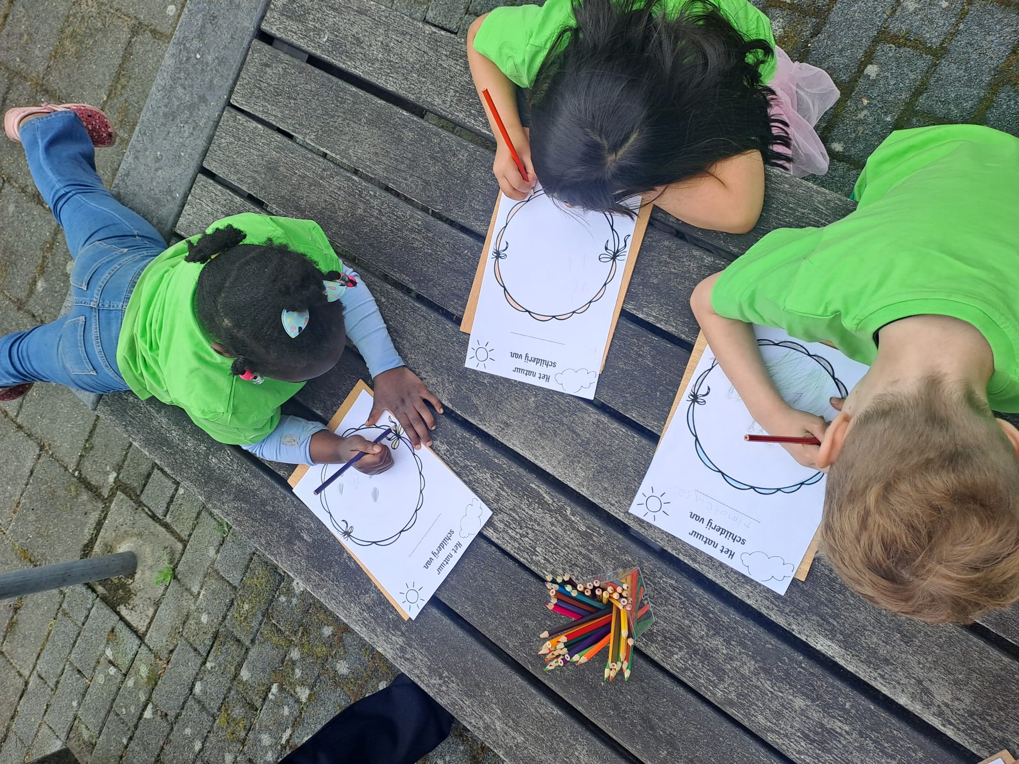 Drie kinderen kleuren en tekenen met potloden op papier aan een houten picknicktafel buiten.