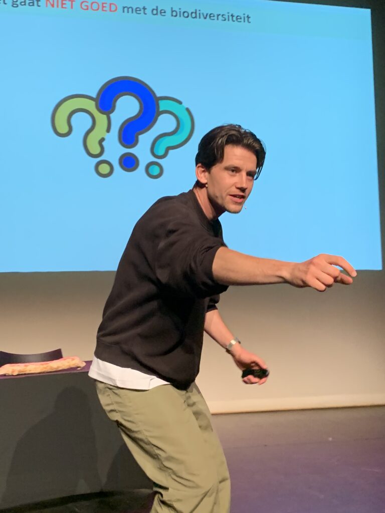 Man presenteert, wijst naar publiek; dia zegt "het gaat NIET GOED met de biodiversiteit" met vraagtekens.