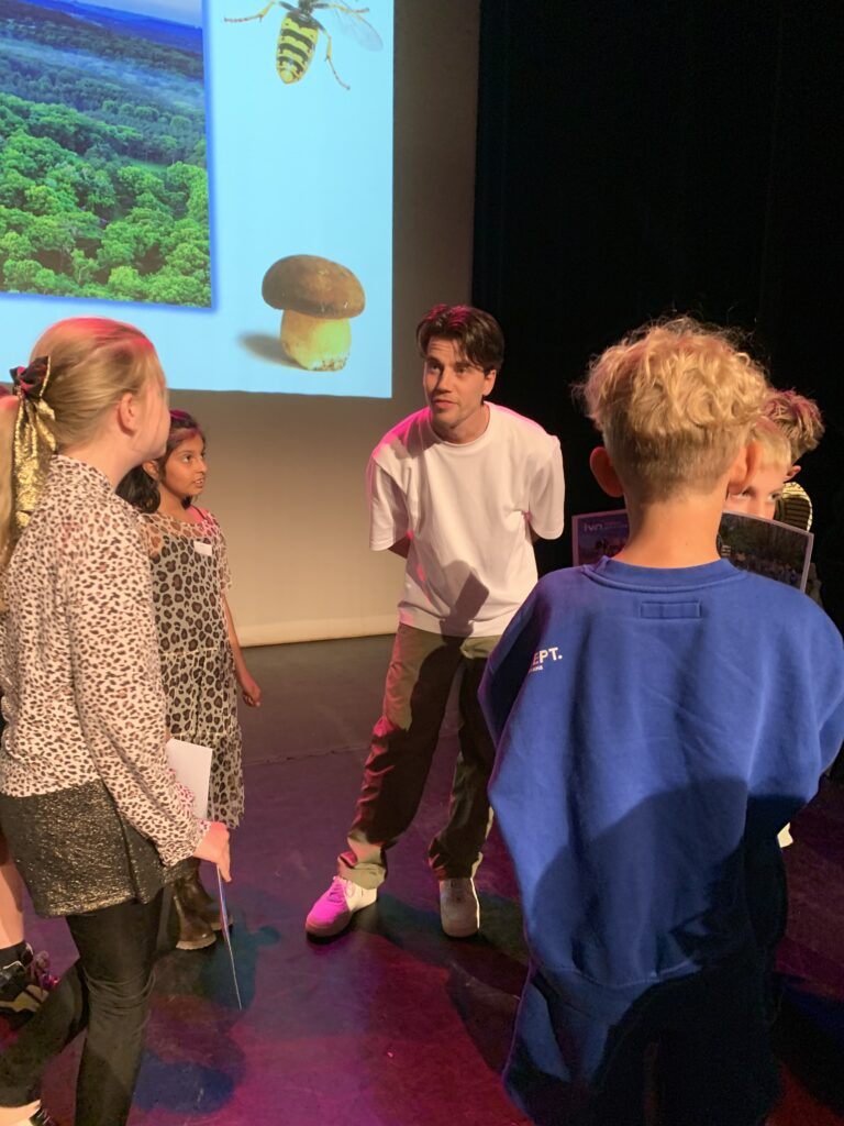 Man in witte shirt praat met kinderen voor een scherm met natuurbeelden en paddenstoel.