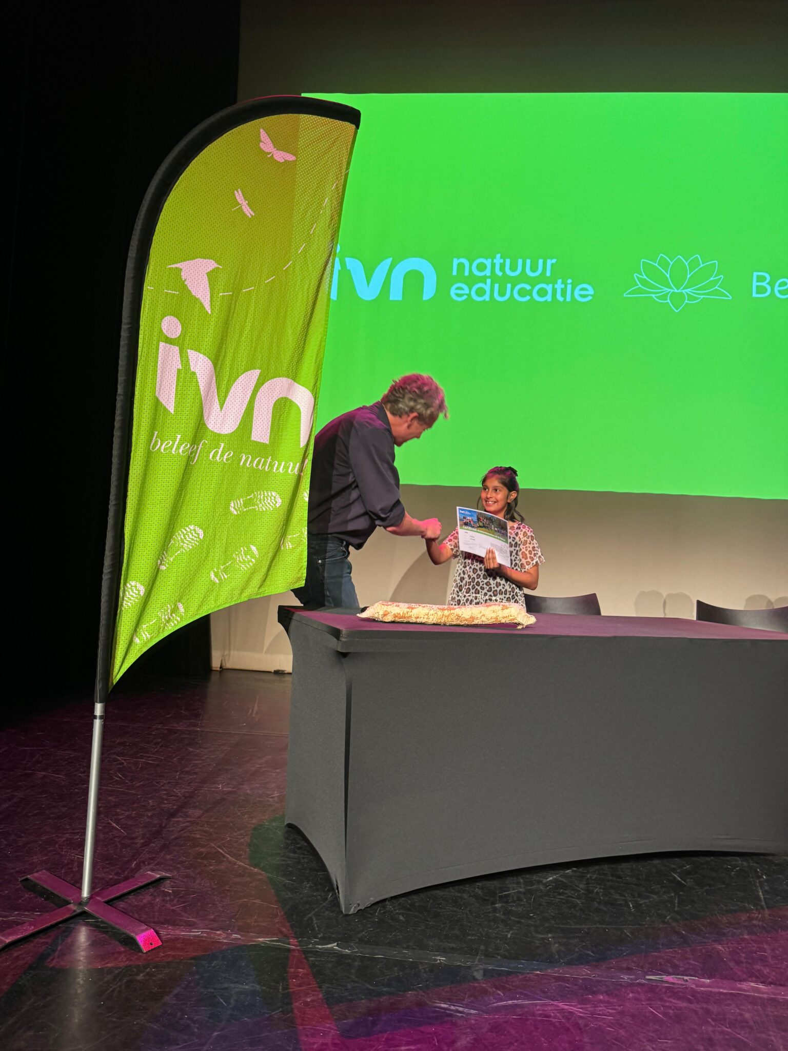 Man en meisje bij groene IVN-banner, met certificaat op podium.