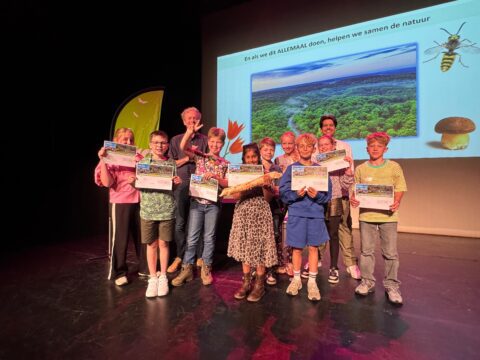 Groep kinderen en een volwassene op podium, houdend certificaten, met natuurafbeelding op scherm.