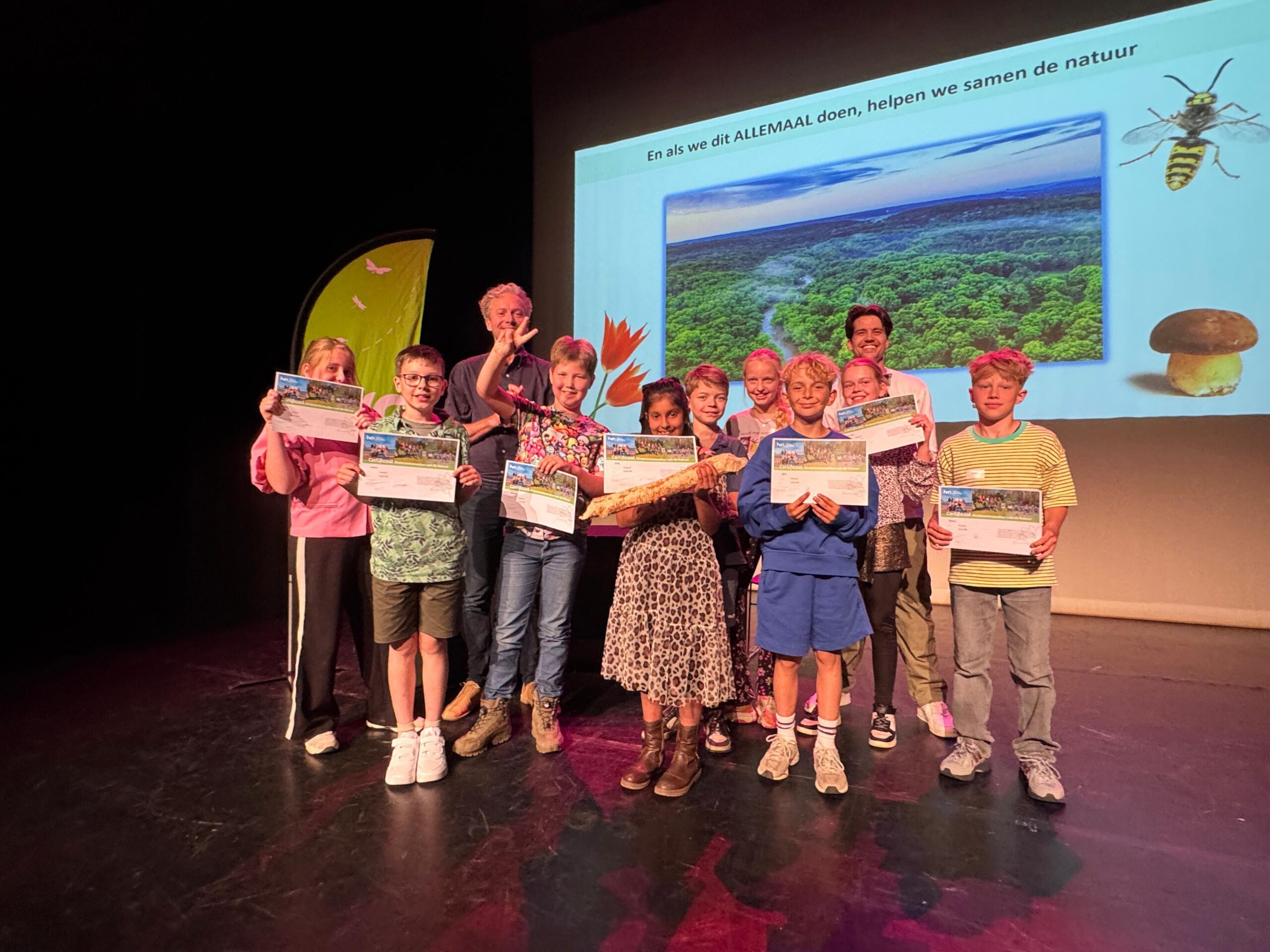 Groep kinderen en een volwassene op podium, houdend certificaten, met natuurafbeelding op scherm.