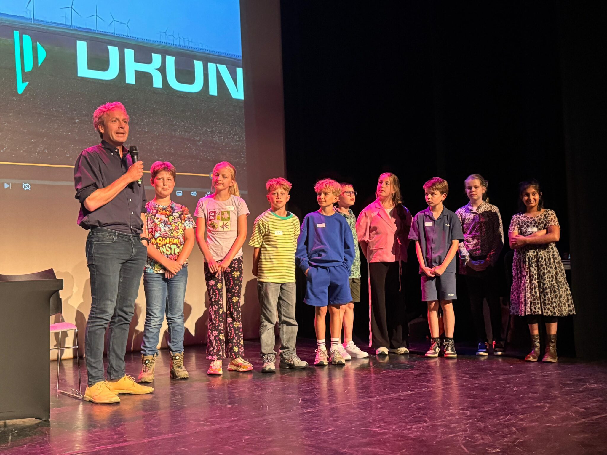 Man spreekt groep kinderen toe op podium met projectiescherm op achtergrond.