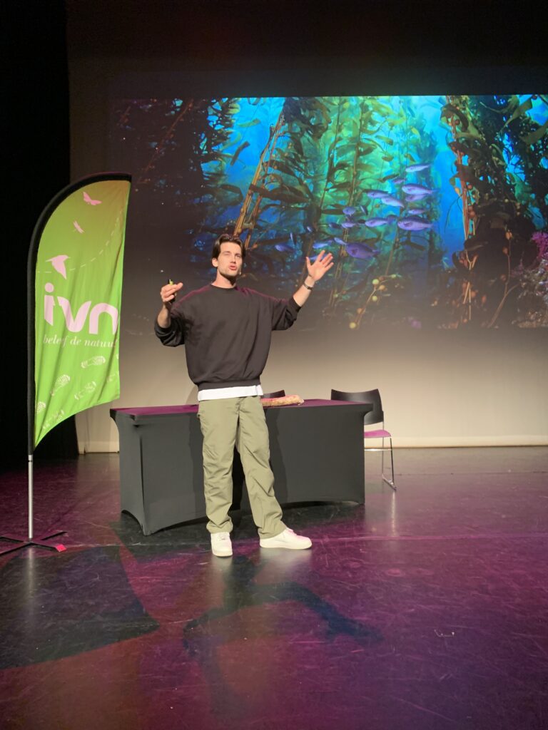 Een man geeft een presentatie voor een onderwaterlandschap met een groen IVN-banner.