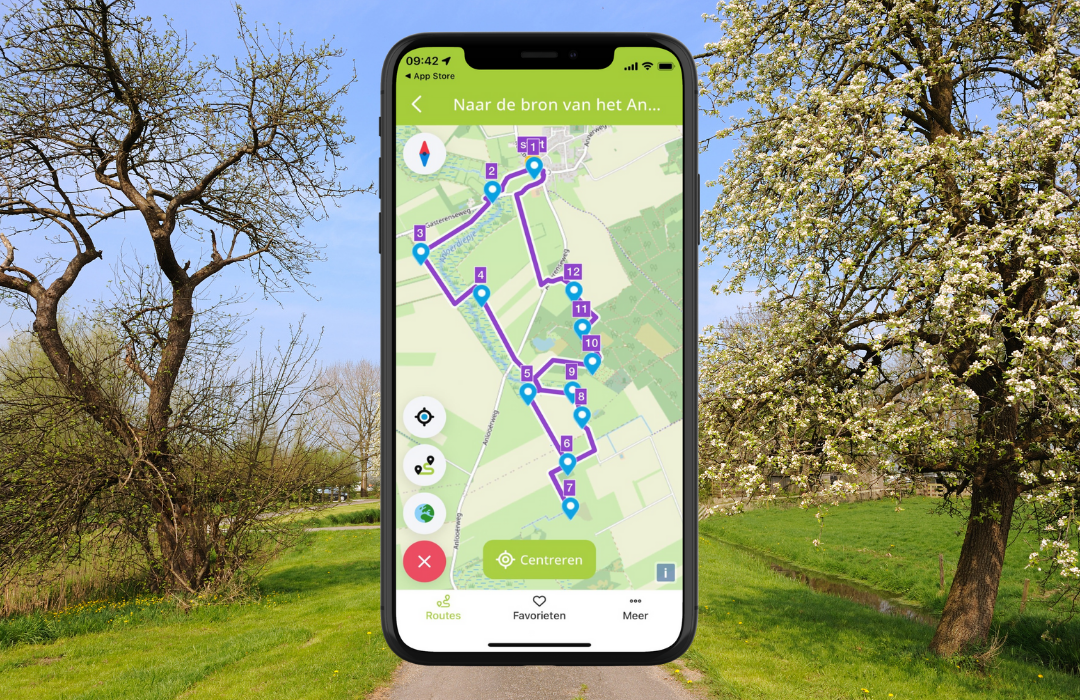 Smartphone toont wandelroutekaart in een groene, landelijke omgeving met bloeiende bomen.