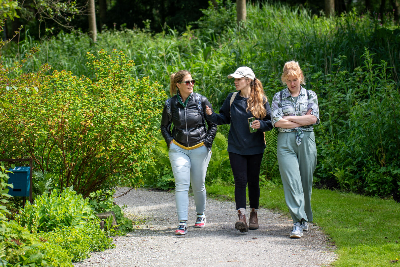 Drie vrouwen wandelen samen op een groen tuinpad, omringd door dichte begroeiing.