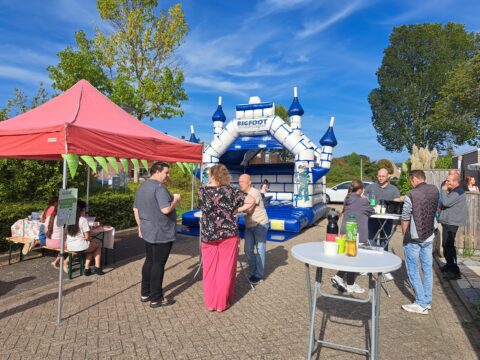 Mensen staan en praten bij een springkasteel en een rode tent op een zonnige dag.