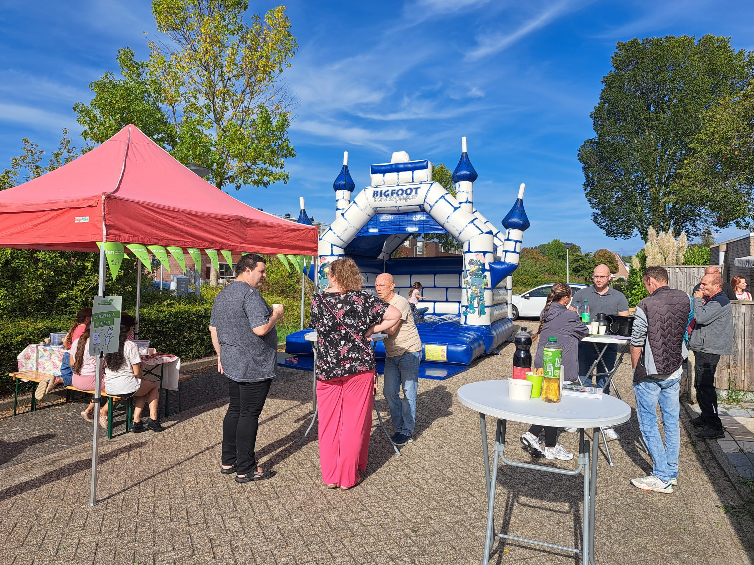 Mensen staan en praten bij een springkasteel en een rode tent op een zonnige dag.