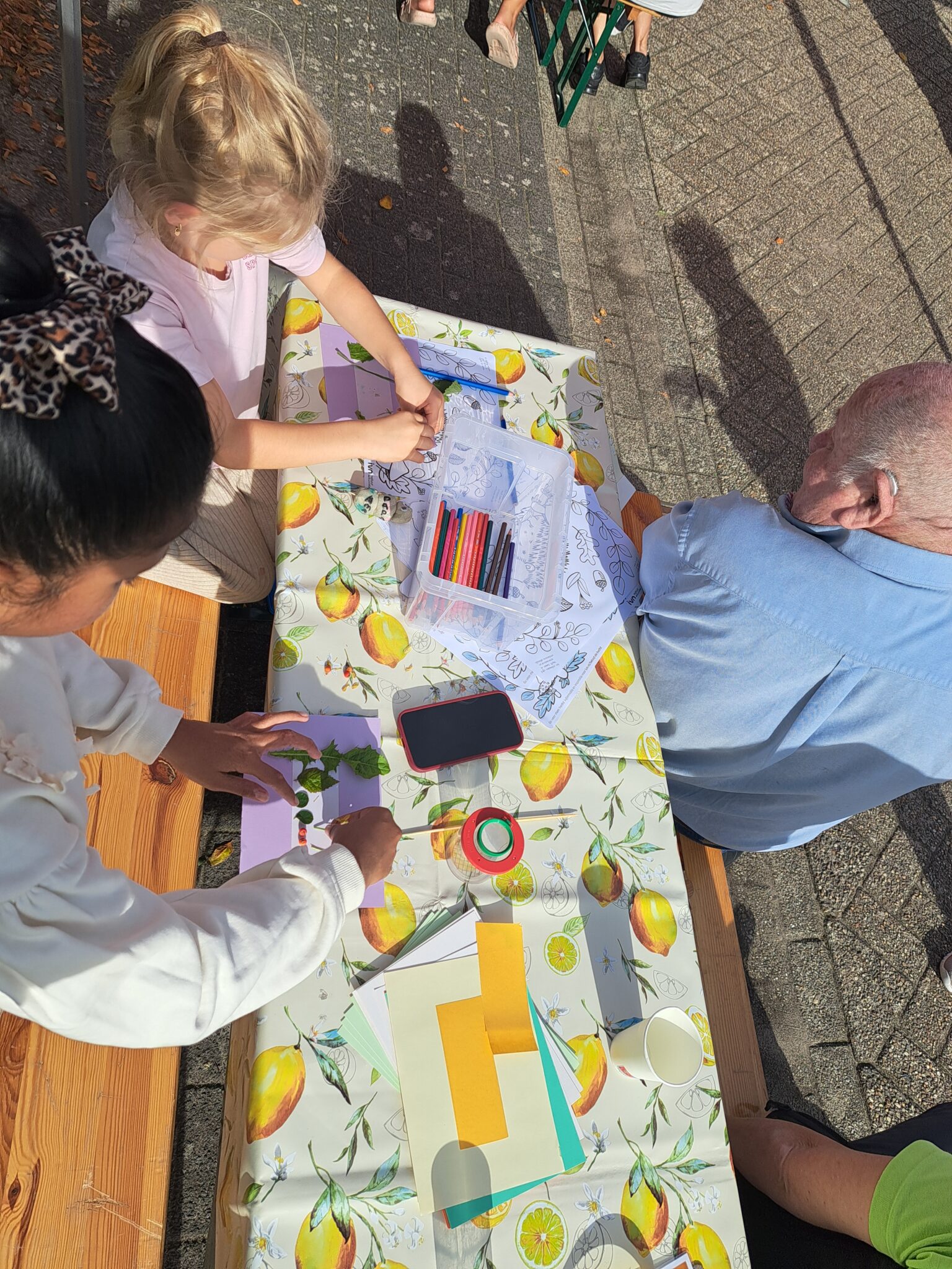 Kinderen knutselen aan een tafel buiten met bladeren, papier en kleurpotloden.