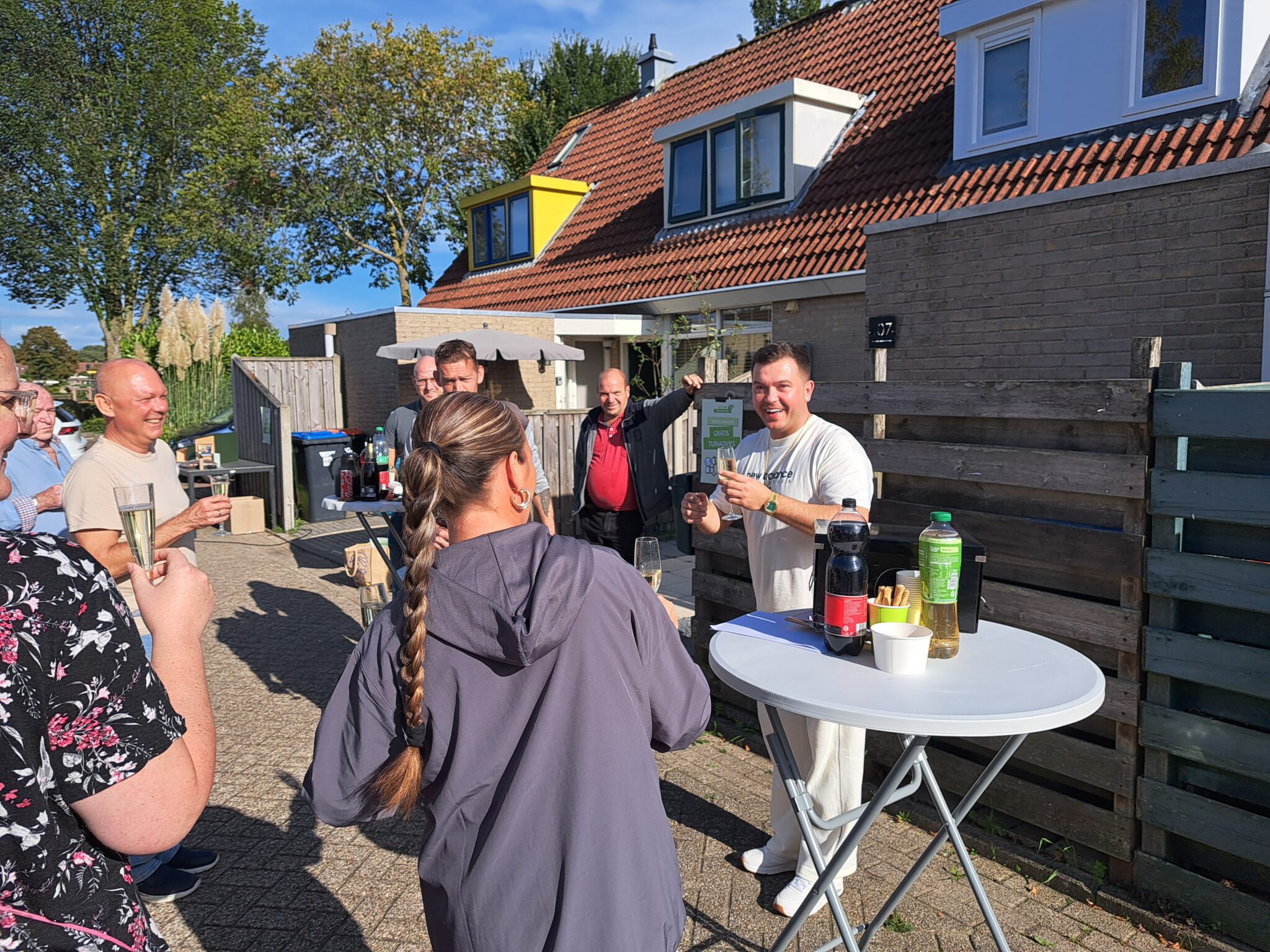 Mensen staan buiten bij een huis, lachend en drinkend rond een statafel met drankjes.