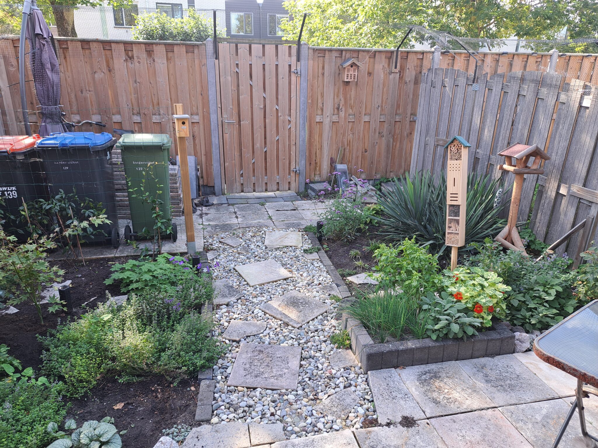Achtertuin met grindpad, plantenborders, vogelhuisjes en kliko's tegen een houten schutting.