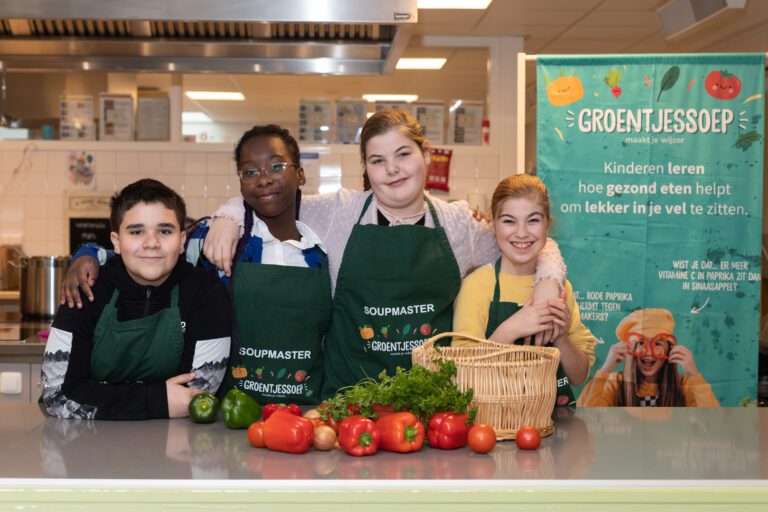 Vier kinderen in groene schorten poseren voor een keuken. Groenten liggen op de toonbank.