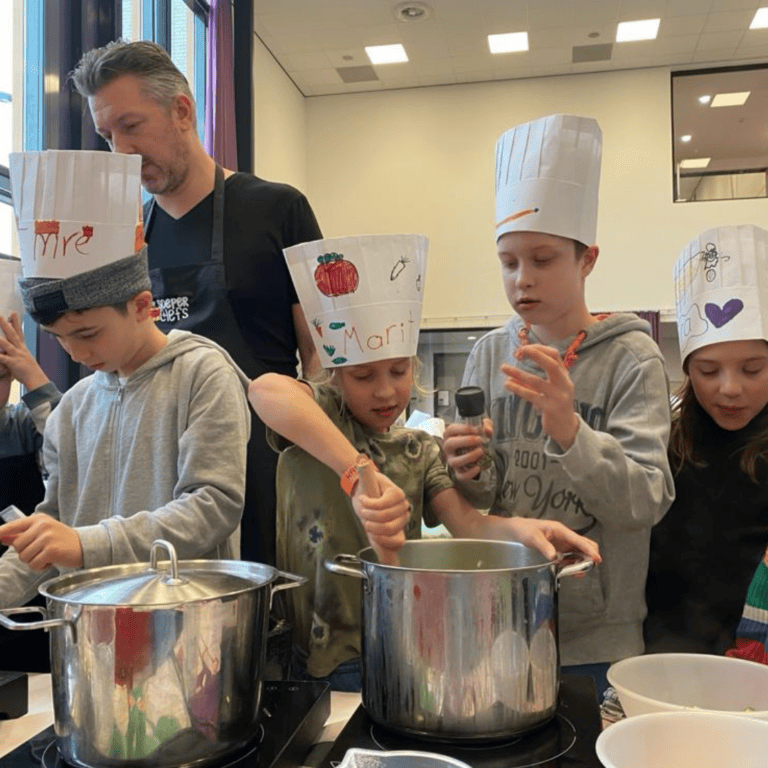 Kinderen met papieren koksmutsen koken samen met een volwassene in een keuken.