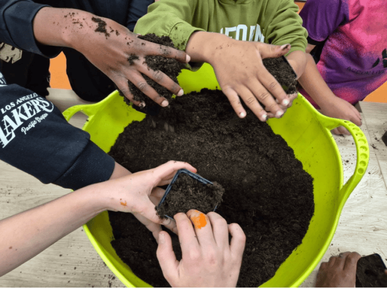 Kinderen vullen plantenpotjes met aarde uit een grote, gele emmer.