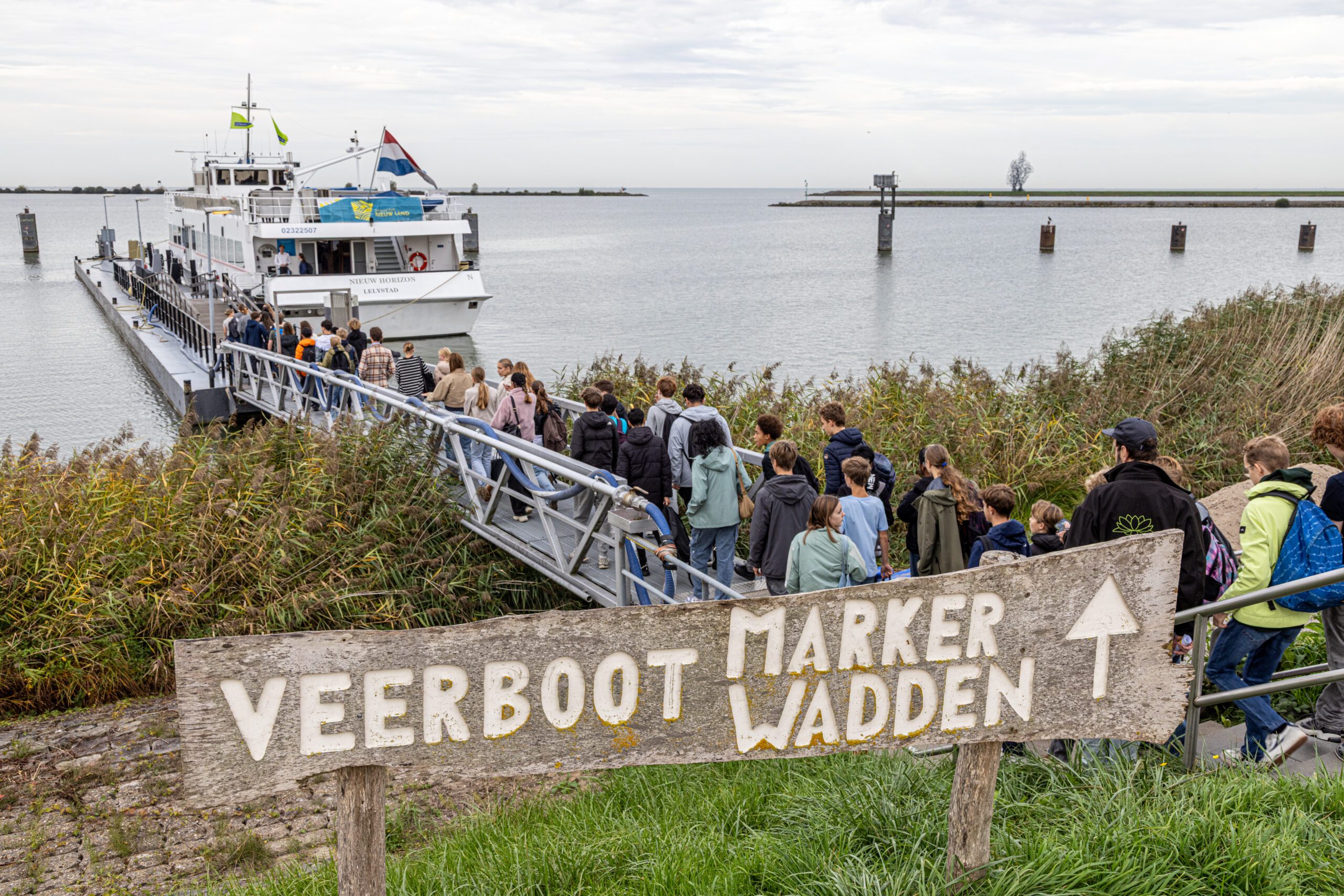 Groep mensen loopt naar een veerboot met een bord "Veerboot Marker Wadden" op de voorgrond.