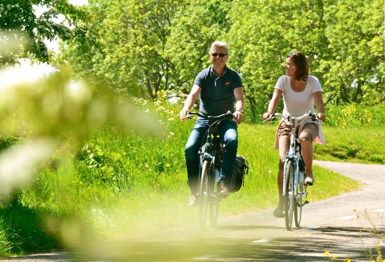 Twee mensen fietsen lachend over een groen, zonnig pad omringd door bomen.
