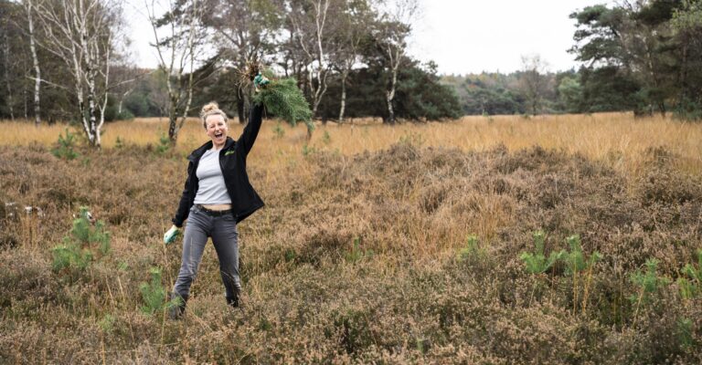 Een vrouw in een veld heft een plant omhoog, omgeven door herfstige natuur en bomen.