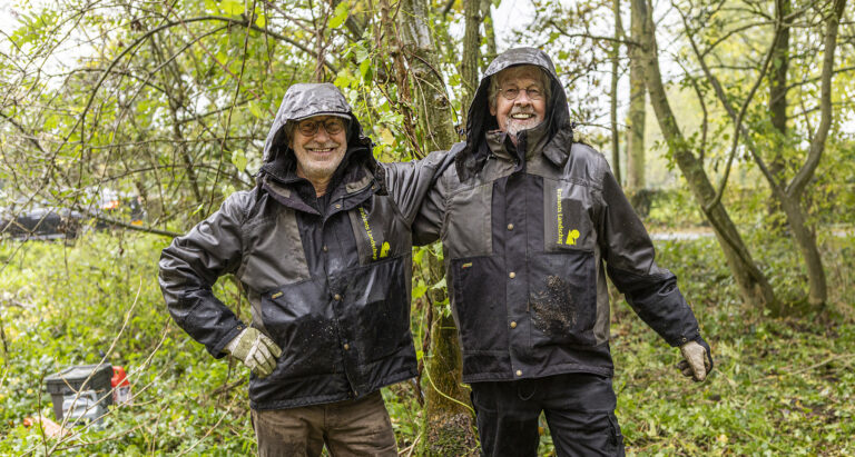 Twee lachende mensen in regenjassen in een bosrijke omgeving omringd door groen.
