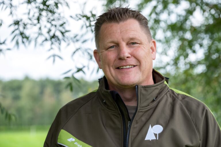 Man in groene jas lacht buiten met groen gebladerte op de achtergrond.