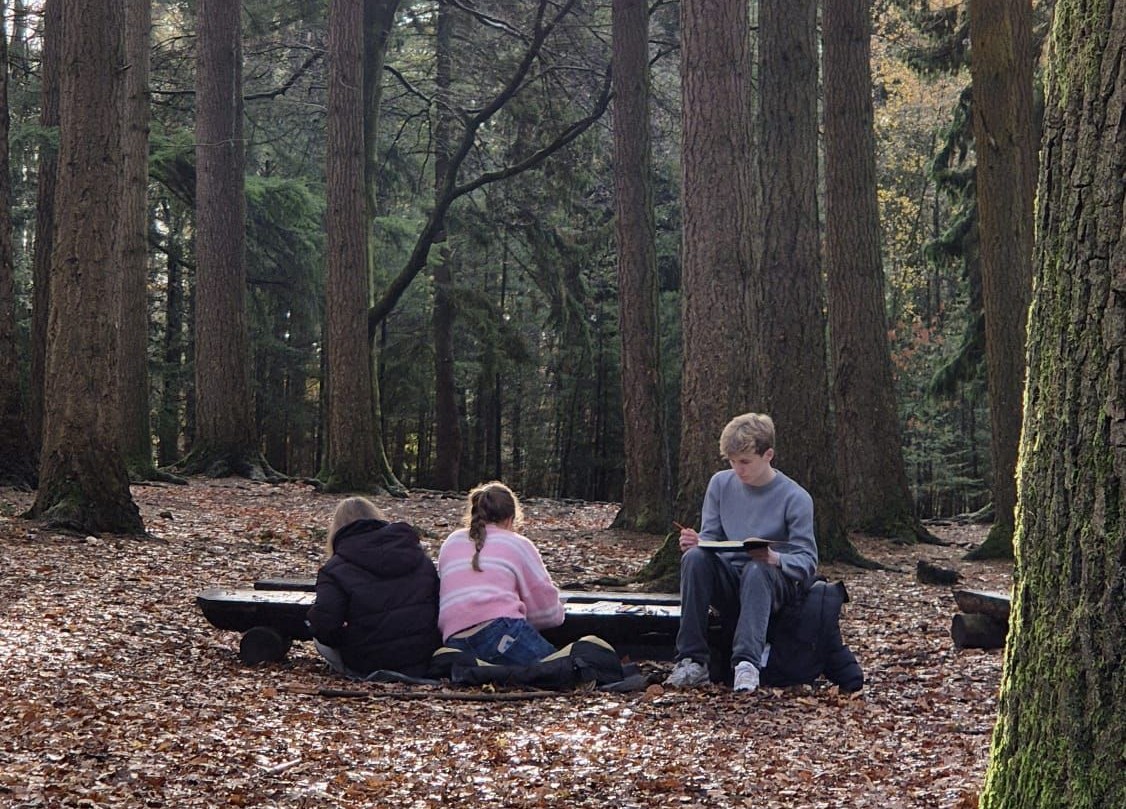 Drie kinderen zitten in een bos op de grond, bezig met boeken en potloden.