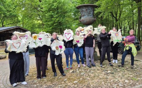 Groep mensen toont versierde stoffen met harten en bloemen, in een bosrijke omgeving.