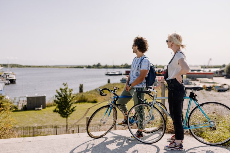 Twee mensen met fietsen kijken over een zonnige rivier met boten en een groene oever.