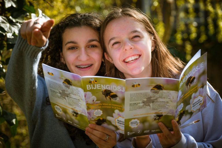 Twee lachende meisjes lezen samen een bijengidsje, terwijl één naar iets in de verte wijst.