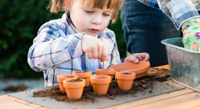 Kind plant zaadjes in kleine terracotta potjes, begeleid door een volwassene in een tuin.