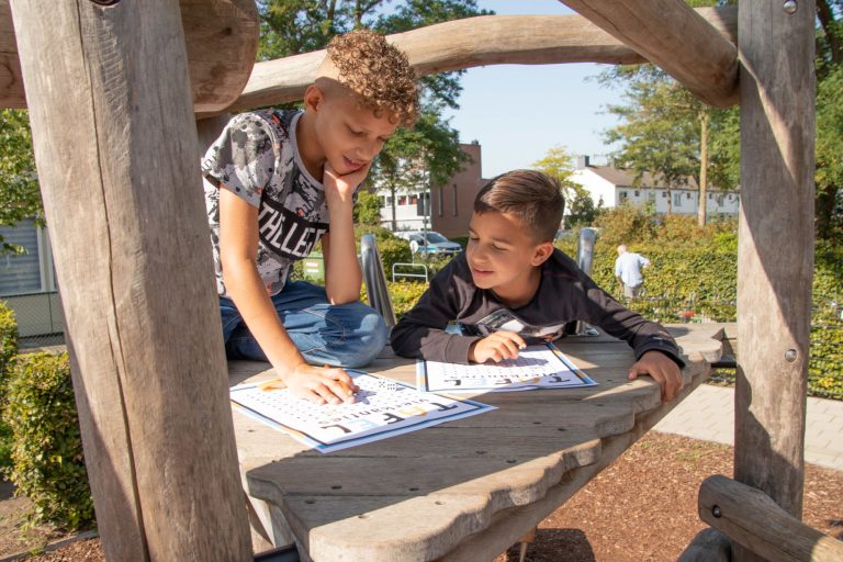 Twee jongens maken woordpuzzels op een houten klimtoestel in een zonnige speeltuin.