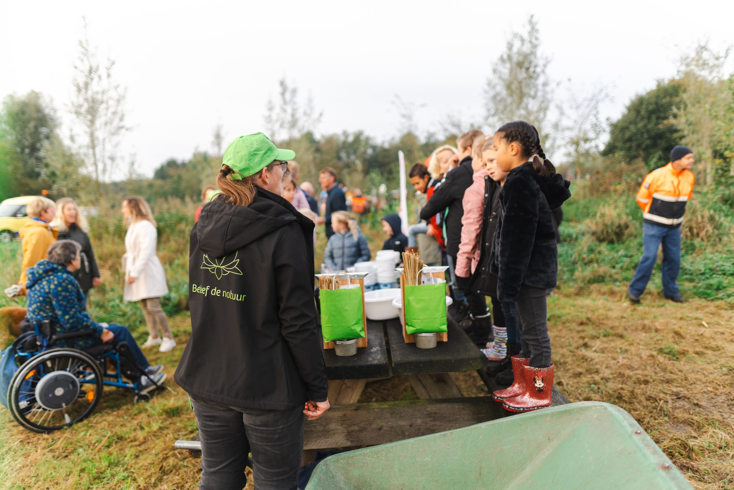 Groep mensen, inclusief kinderen, verzamelen in een natuurgebied bij een tafel met groene tassen.