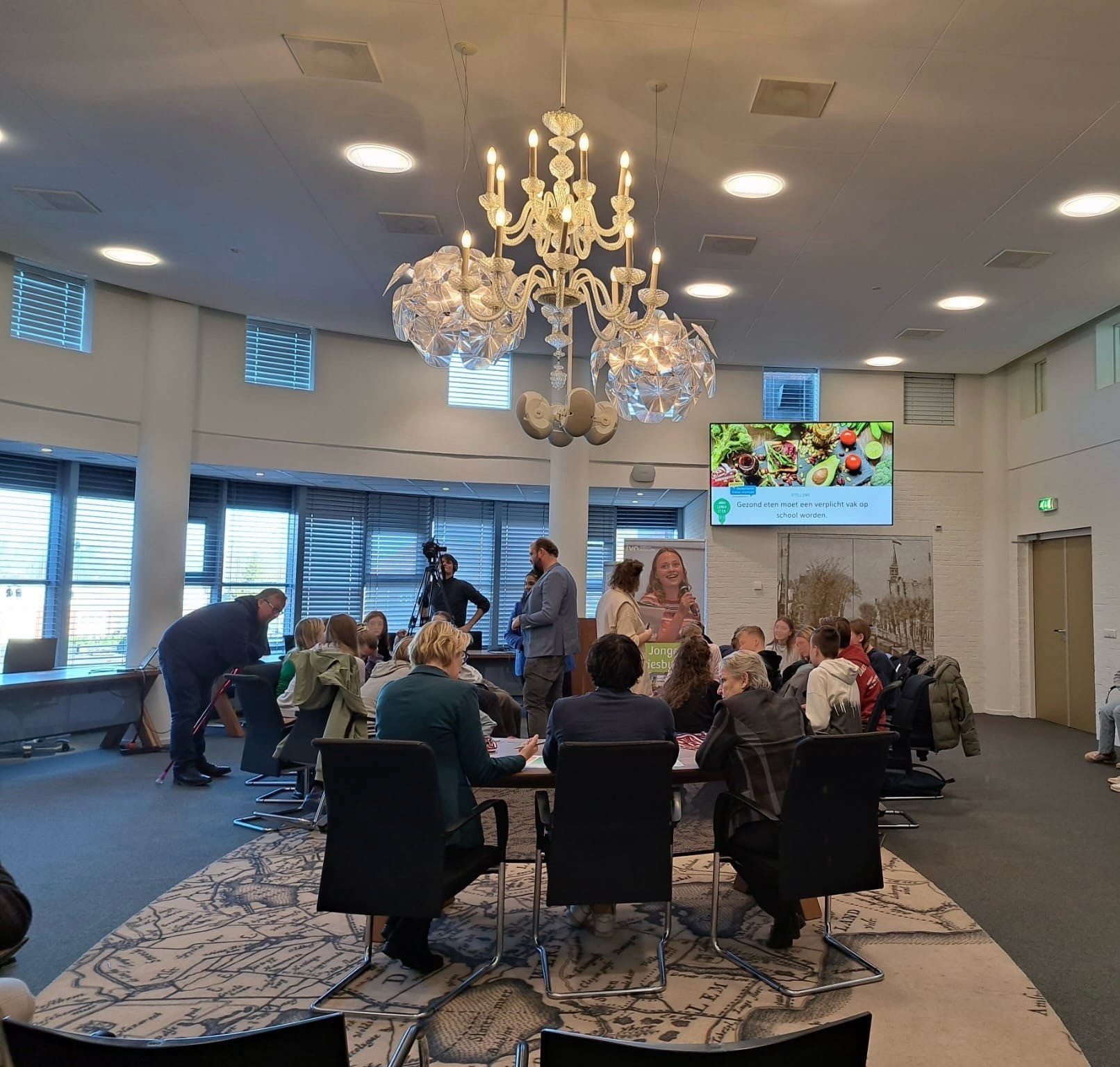 Vergaderruimte met mensen rond tafel, scherm toont presentatie over gezond eten op school.