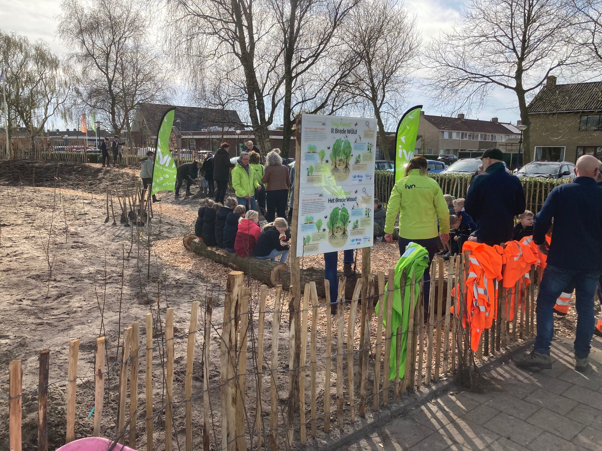 Mensen verzamelen zich rond borden en vlaggen bij een omheind gebied met jonge boompjes.