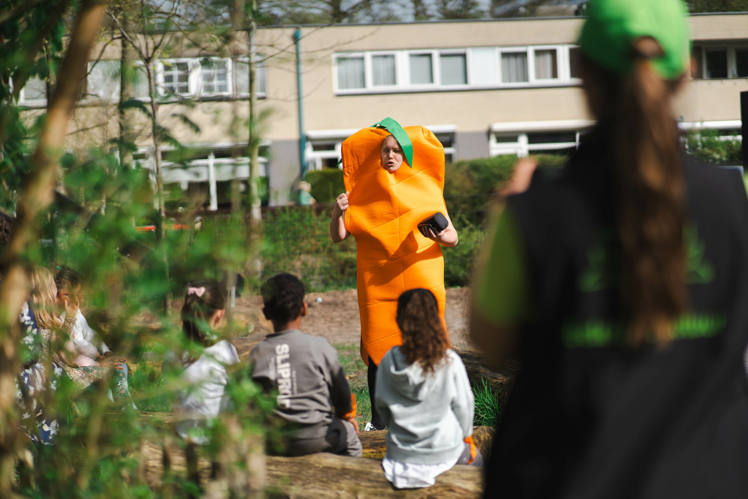 Persoon verkleed als wortel spreekt kinderen toe in een tuinachtige omgeving met schoolgebouw op achtergrond.