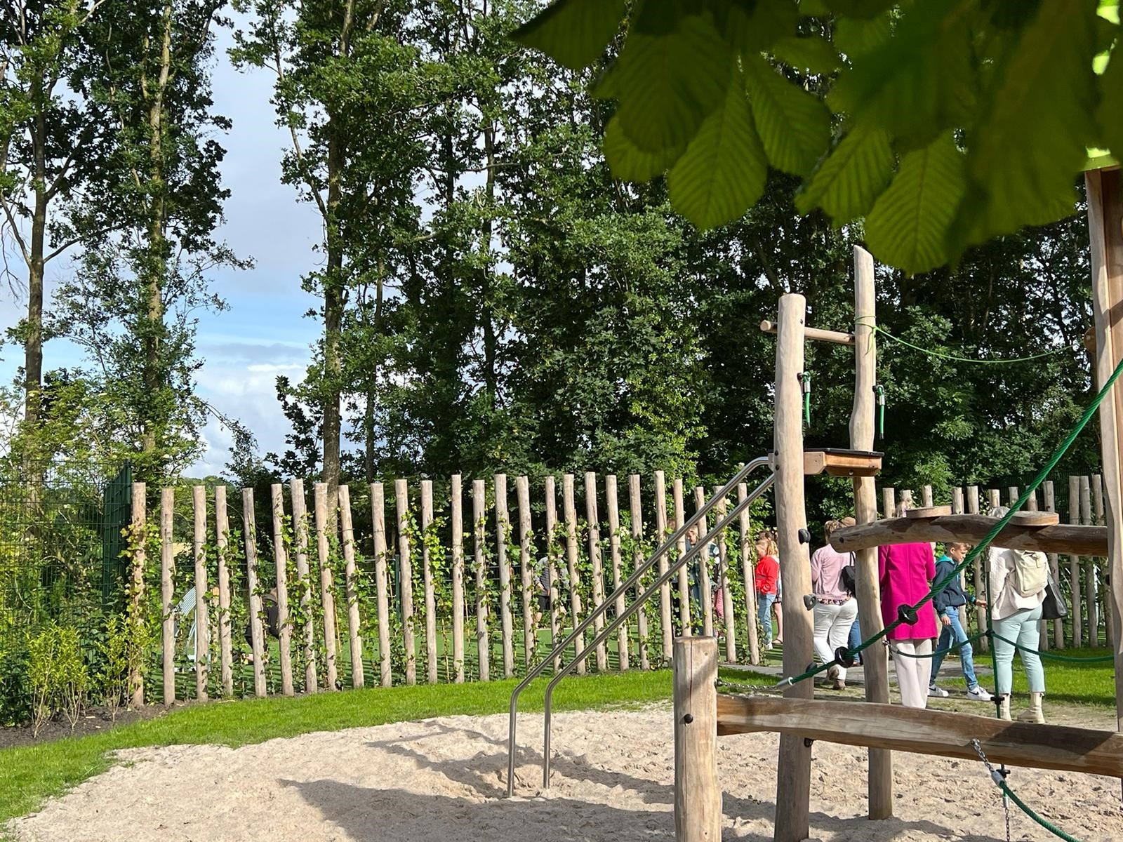 Speeltuin met houten klimtoestel, omringd door bomen en schutting, enkele mensen op de achtergrond.