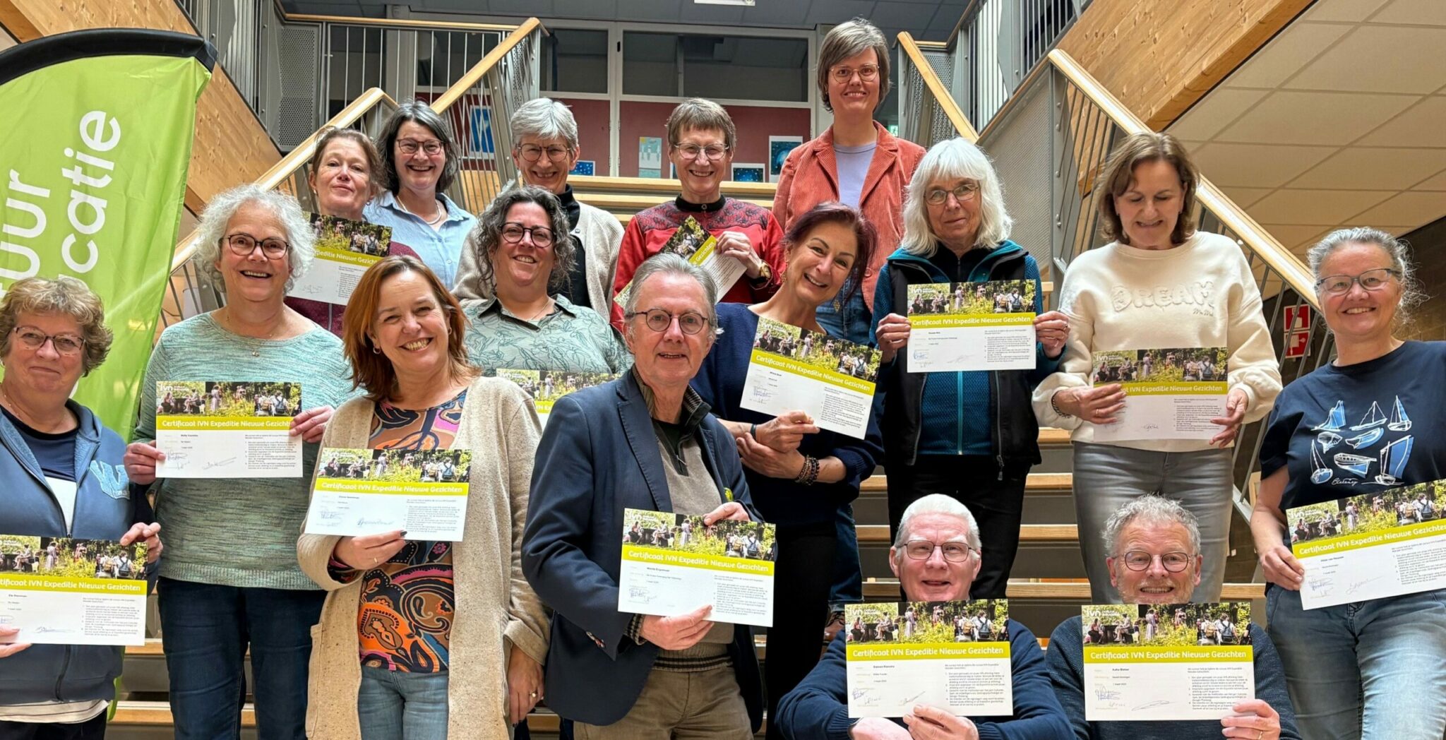 Groep volwassen mensen op een trap met certificaten, groene banner links.