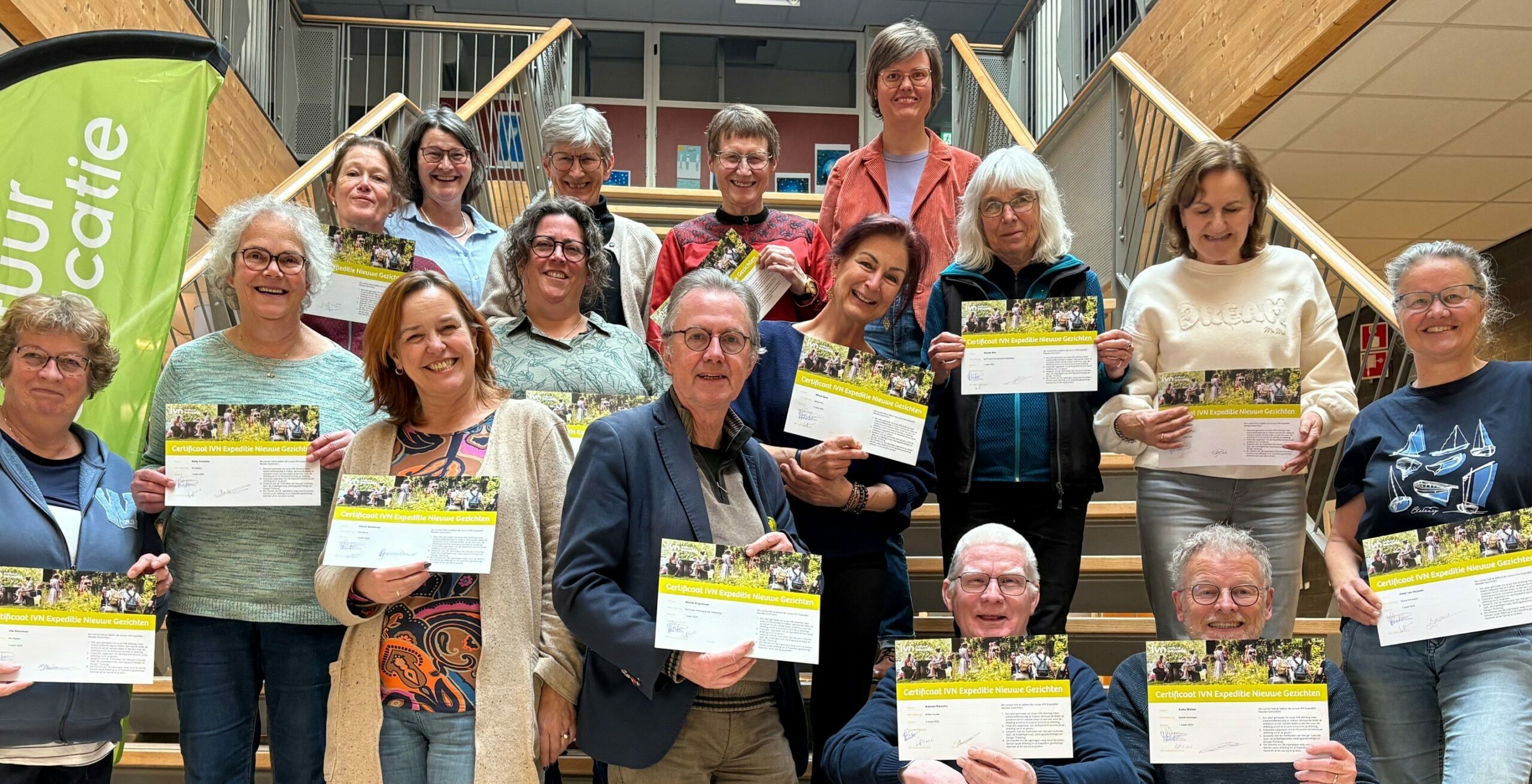 Groep volwassen mensen op een trap met certificaten, groene banner links.