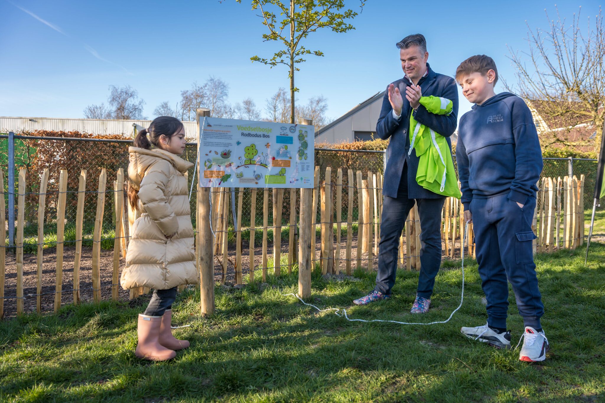 Kinderen en een man bij een informatiebord in een tuin, met houten omheining en gras.