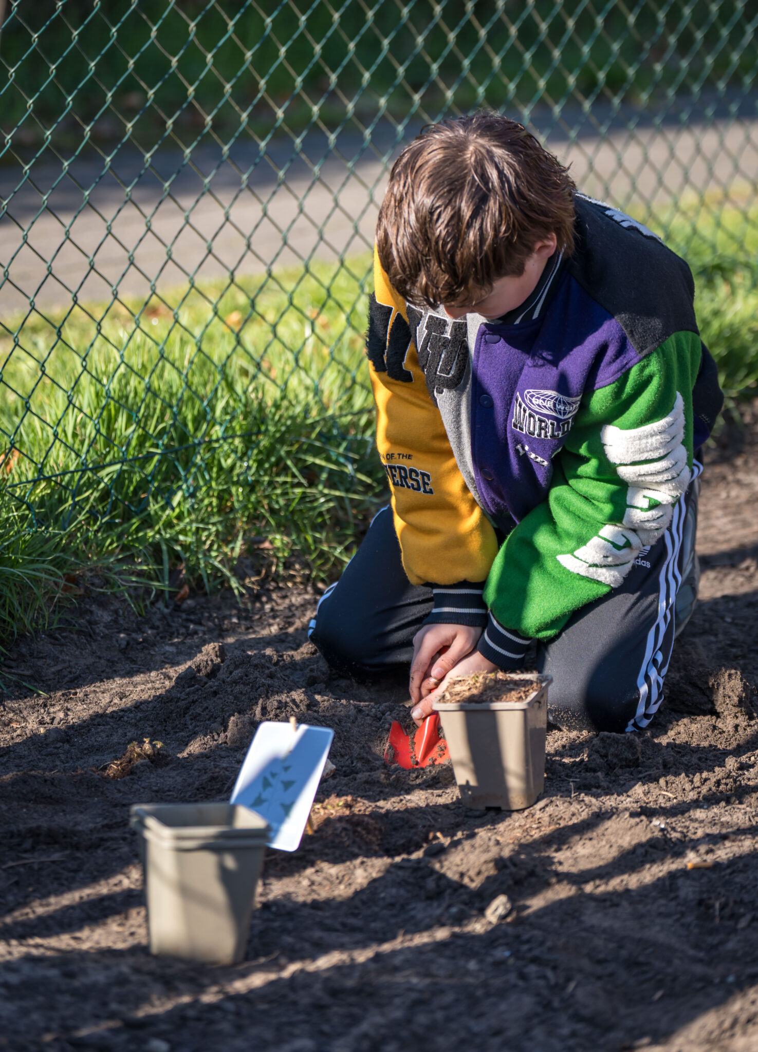 Een kind plant in een tuin, gehurkt bij een hek met plantpotten en labels op de grond.