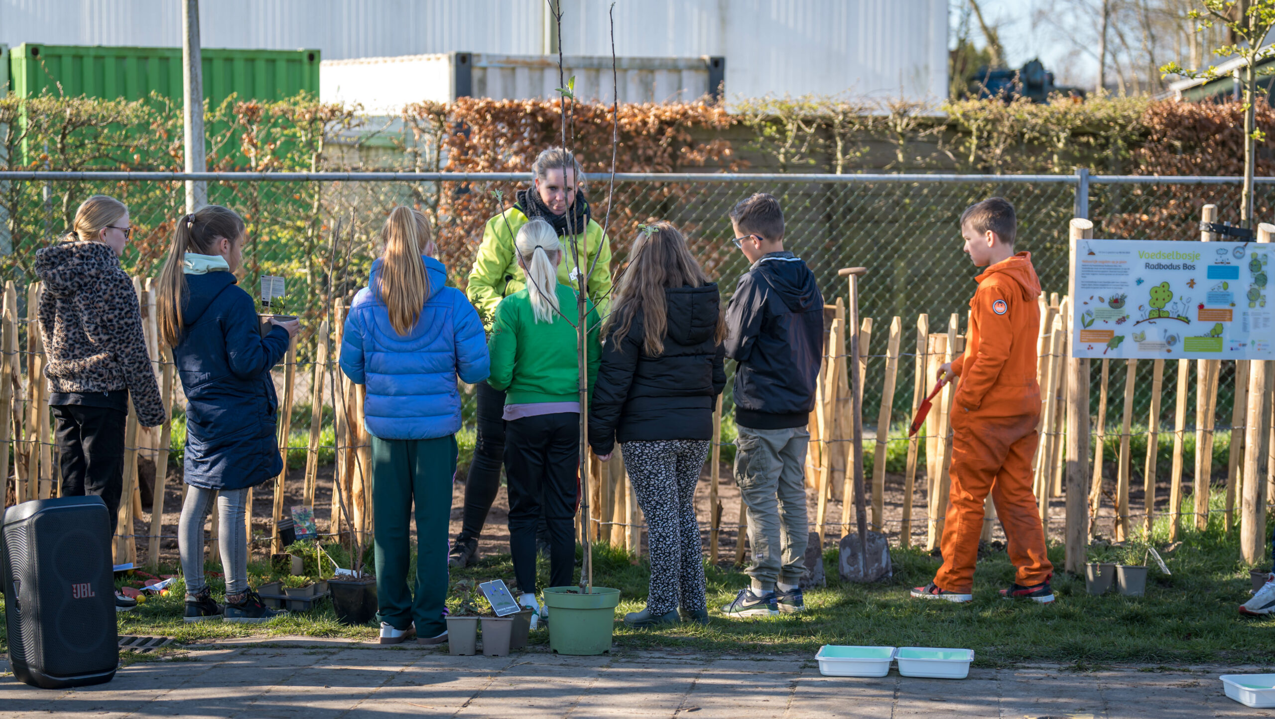 Kinderen planten bomen in een tuin, begeleid door een volwassene, bij een hek met een poster.