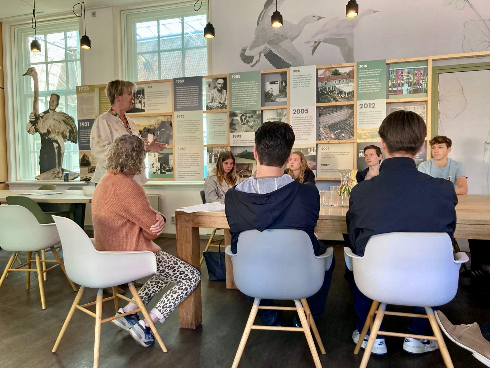 Groep mensen in een lesruimte, docent staat voor een wand met historische posters.