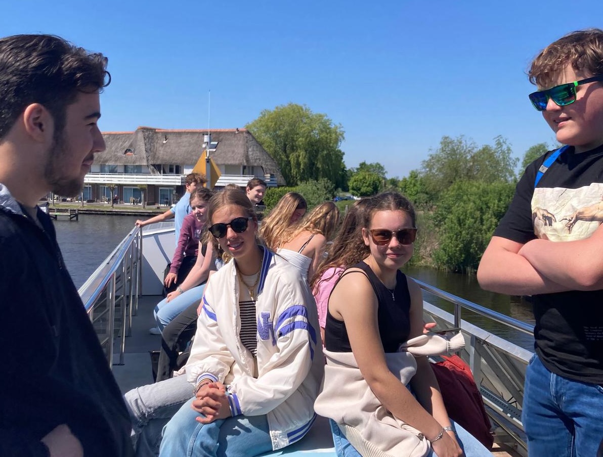 Groepje jongeren op een boot, zonnige dag, rietgedekte huizen en groen op de achtergrond.
