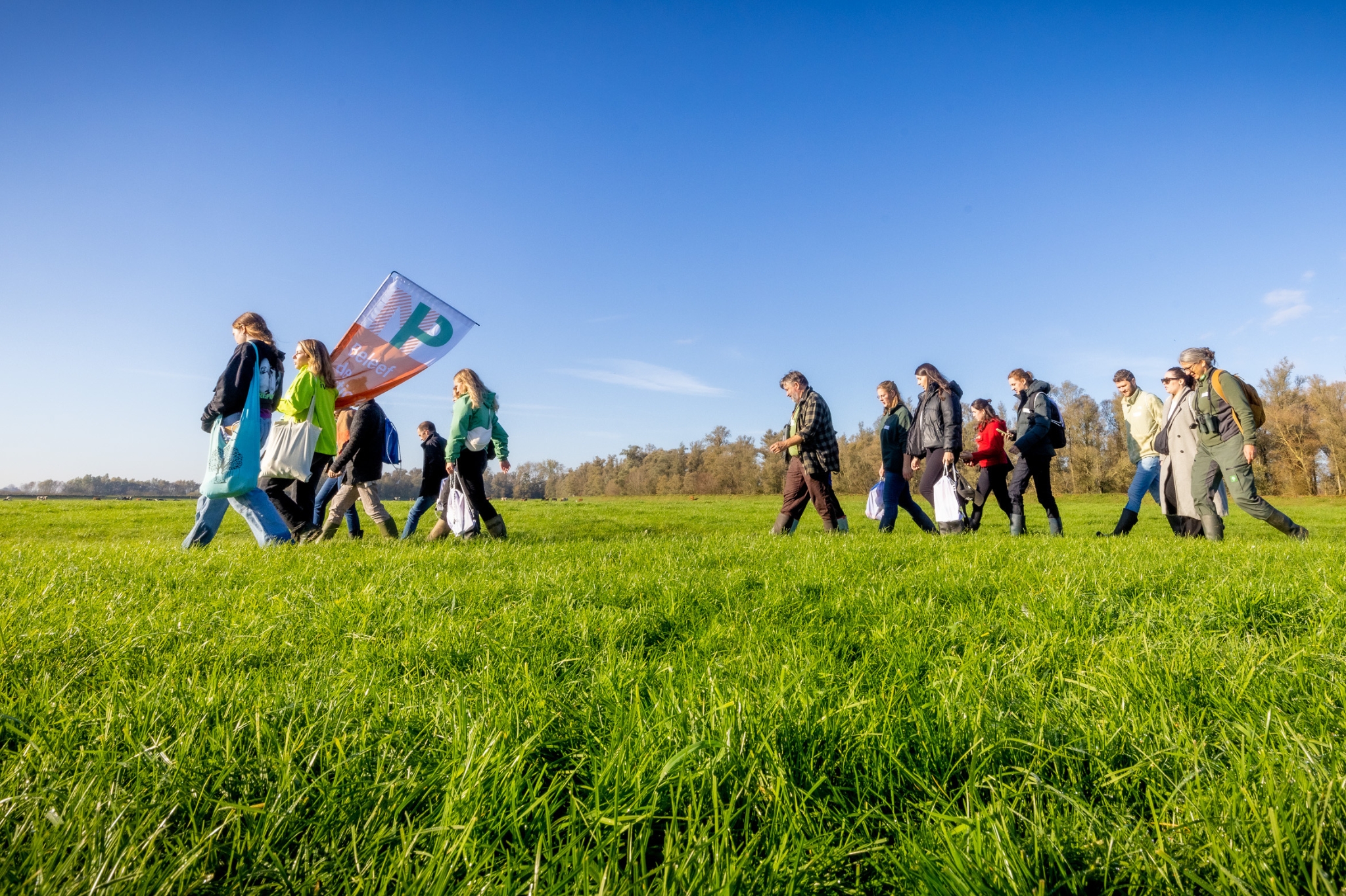 Een groep wandelaars met een vlag loopt over een groene weide onder een heldere blauwe lucht.