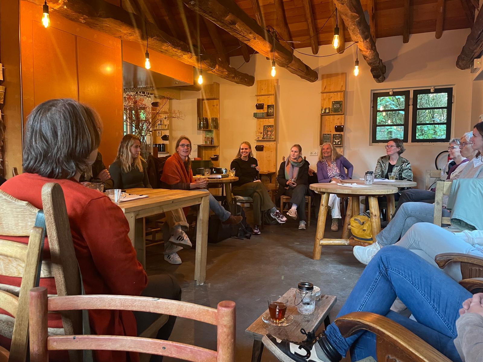 Groep mensen zit in een kring binnen, gezellig onder houten balken en lamplicht.