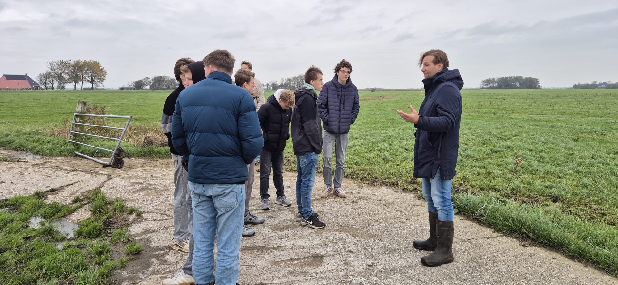 Mensen praten op een boerderijerf naast een weiland, onder een bewolkte hemel.