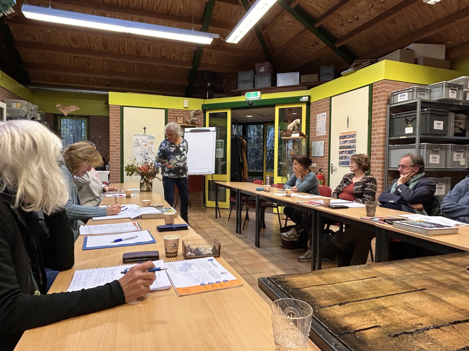 Groep mensen in vergaderruimte, luisteren naar spreker bij flip-over. Tafels met documenten.