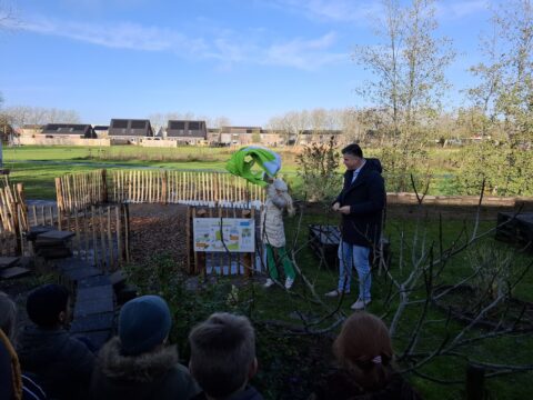 Twee mensen onthullen een bord in een tuin met toeschouwers, huizen op de achtergrond.