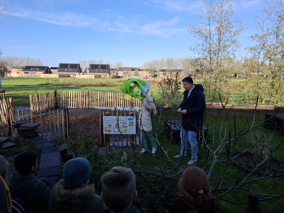 Twee mensen onthullen een bord in een tuin met toeschouwers, huizen op de achtergrond.